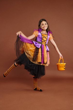 cheerful girl in Halloween costume holding bucket with candies and holding skirt on brown backdrop