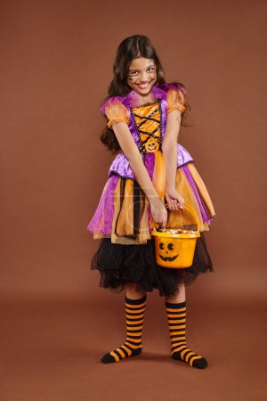 delightful girl in Halloween costume holding bucket with candies and holding skirt on brown backdrop