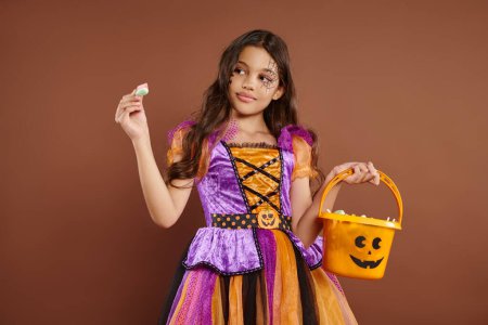 niedliches Mädchen im Halloween-Kostüm mit Eimer und Blick auf verpackte Bonbons auf braunem Hintergrund