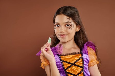cheerful girl in Halloween costume holding sweet jelly candy on brown backdrop, trick or treat