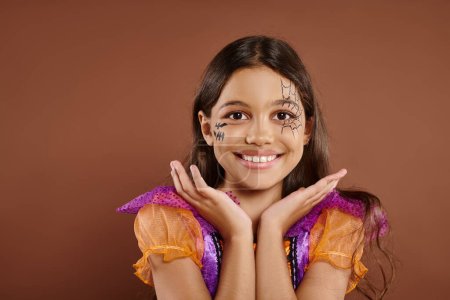 fröhliches Mädchen im Halloween-Kostüm und Spinnennetz-Make-up gestikulierend auf braunem Hintergrund, Trick oder Leckerbissen