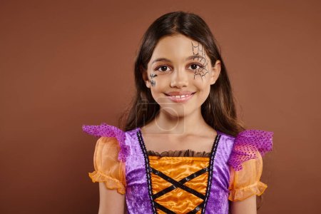 adorable girl in Halloween costume and spiderweb makeup smiling on brown backdrop, trick or treat