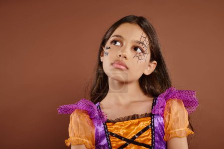 nachdenkliches Mädchen in buntem Kostüm mit Halloween-Make-up auf braunem Hintergrund, Oktober