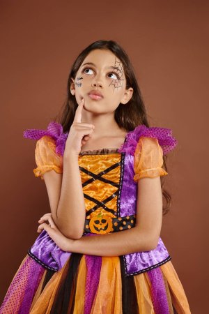 thoughtful girl in colorful costume with Halloween makeup looking away on brown background, October