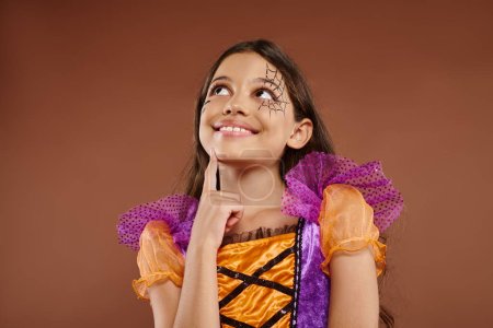 Photo pour Fille rêveuse en costume coloré avec maquillage Halloween regardant loin sur fond brun, visage heureux - image libre de droit