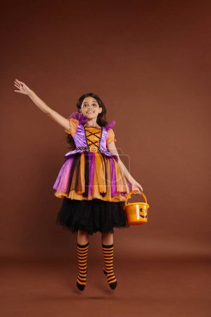 happy girl in Halloween costume levitating with candy bucket on brown background, magic concept