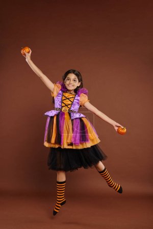 happy girl in Halloween costume levitating with pumpkins on brown background, magic concept