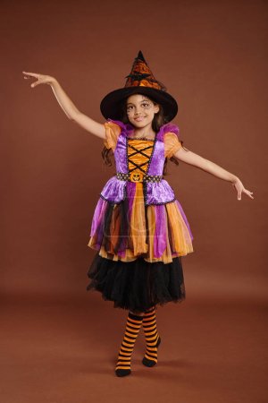 anmutiges Mädchen im Halloween-Kostüm und Hexenhut posiert auf braunem Hintergrund, magisches Konzept