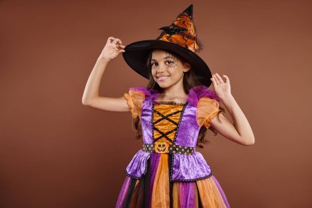 happy girl in Halloween costume and pointed hat posing on brown background, little witch attire