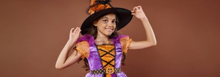 chica feliz en disfraz de Halloween y sombrero puntiagudo posando sobre fondo marrón, pequeña bandera de bruja
