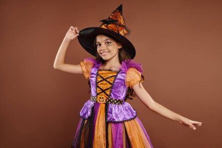 chica alegre en disfraz de Halloween y sombrero puntiagudo posando sobre fondo marrón, pequeño atuendo de bruja
