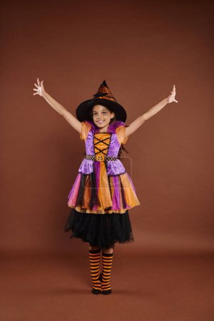 excited girl in Halloween costume and pointed hat standing with raised hands on brown background