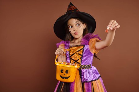 chica divertida en disfraz de Halloween y sombrero puntiagudo sosteniendo cubo y mostrando dulces sobre fondo marrón