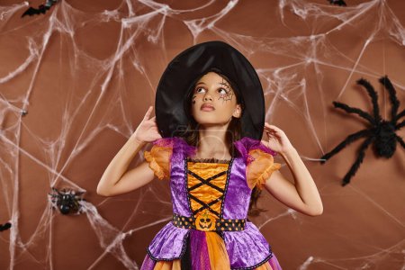 Mädchen im Halloween-Kostüm und mit Hexenhut, die nach oben schaut und neben Spinnweben auf braunem Hintergrund steht