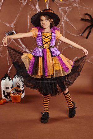 girl in witch hat and dress holding skirt near bucket with sweets on brown, Halloween concept