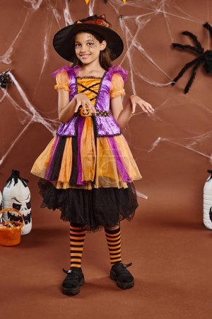fröhliches Mädchen mit Hexenhut und Kleid tanzt in der Nähe von Eimer mit Süßigkeiten auf braun, Halloween-Konzept