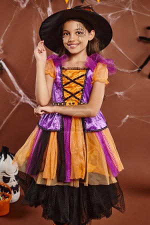 positive girl in witch costume and pointed hat smiling on brown background, Halloween concept