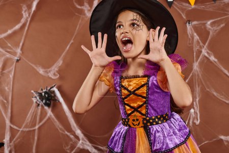Emotionales Mädchen mit Hexenhut und Halloween-Kostüm schreit und gestikuliert auf braunem Hintergrund
