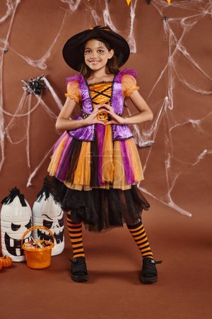 happy little witch in Halloween costume and pointed hat showing heart gesture on brown backdrop
