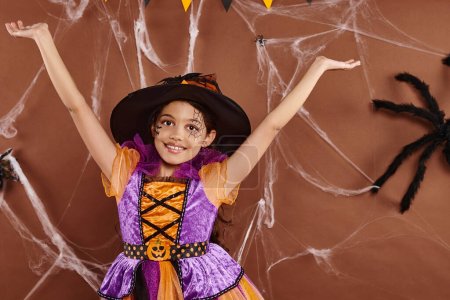 joyous little witch in Halloween costume and pointed hat with raised hands on brown backdrop