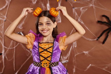 chica divertida con maquillaje de tela de araña sonriendo y sosteniendo calabazas cerca de la cabeza en el fondo marrón, Halloween