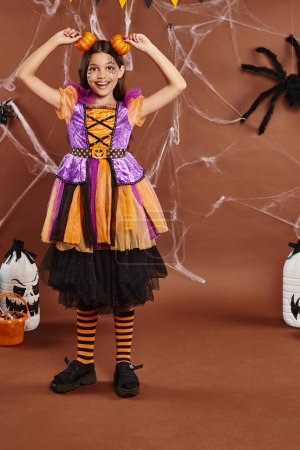 cheerful girl in Halloween bright dress holding pumpkins near head on brown backdrop, spooky season