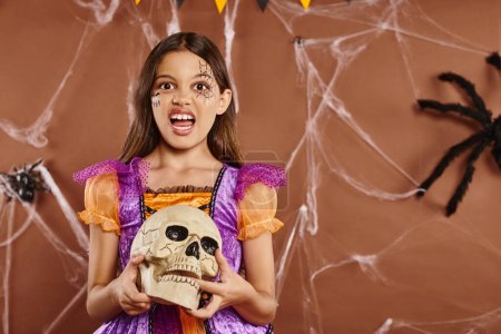 Foto de Chica en disfraz de Halloween sosteniendo el cráneo y gruñendo en la cámara sobre fondo marrón, temporada espeluznante - Imagen libre de derechos