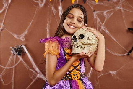 Photo for Delightful girl in dress holding skull and smiling on brown background, Halloween spooky season - Royalty Free Image