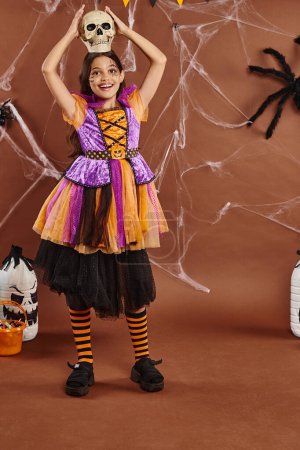 Photo for Joyful girl in Halloween dress standing with skull on head on brown background, spooky season - Royalty Free Image