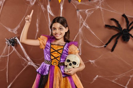 fröhliches Mädchen im Halloween-Kleid steht mit Totenkopf und zeigt mit dem Finger nach oben auf braunem Hintergrund