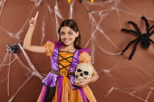 cheerful girl in Halloween dress standing with skull and pointing up with finger on brown background t-shirt #676715740
