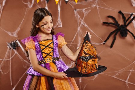 happy girl in Halloween witch costume holding pointed hat on brown background, spooky season