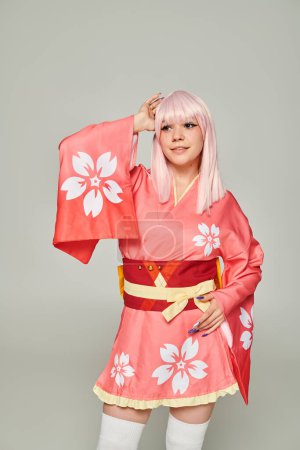 Foto de Joven mujer sonriente en peluca rubia y kimono rosa con estampado floral en gris, estilo anime - Imagen libre de derechos