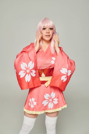 young blonde woman with colorful manicure wearing pink kimono and looking at camera on grey