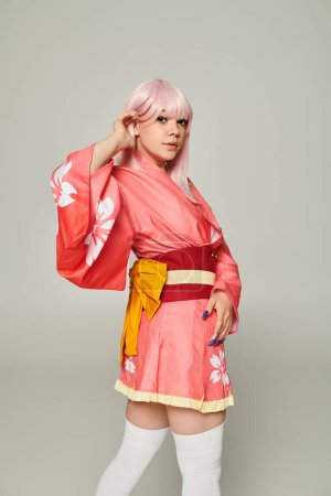 charming woman in colorful japanese style attire adjusting blonde hair and looking at camera on grey