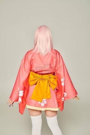vista trasera de la mujer en peluca rubia y kimono rosa corto con lazo amarillo de pie en gris, estilo anime