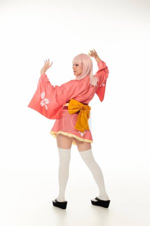 Foto de Mujer joven estilo anime en kimono rosa con lazo amarillo y calcetines blancos de rodilla en blanco, longitud completa - Imagen libre de derechos