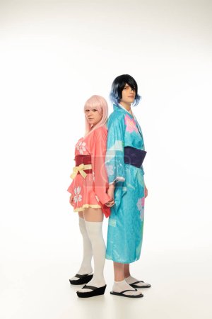 young cosplayers in colorful kimonos and wigs standing back to back and holding hands on white