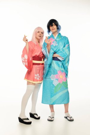 full length of young anime couple in bright kimonos showing mini hearts signs with fingers on white