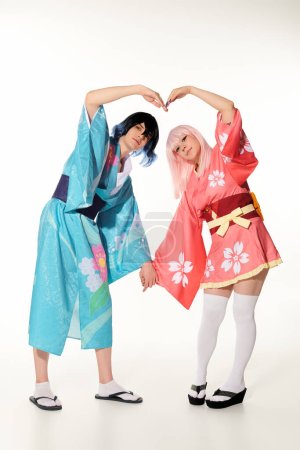 Téléchargez les photos : Cosplayers romantiques en perruques et kimonos lumineux montrant grand signe du cœur avec les mains sur blanc - en image libre de droit