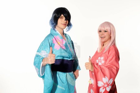anime couple in wigs and kimonos showing thumbs up and looking at camera on white, cosplay trend