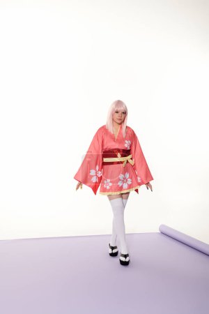 junge Frau im Anime-Stil in rosa Kimono und blonder Perücke auf lila Teppich und weißem Hintergrund, Cosplay