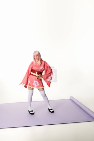mujer de estilo anime en kimono rosa y peluca rubia con la mano en la cadera en la alfombra púrpura en estudio blanco