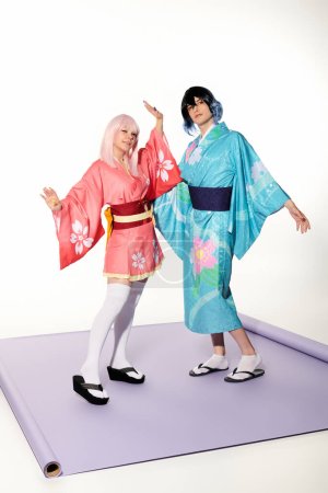 junge ausdrucksstarke Cosplayer in bunten Kimonos und Perücken posieren auf lila Teppich im weißen Studio