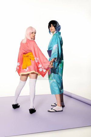 anime estilo pareja en kimonos cogidas de la mano y mirando a la cámara en la alfombra púrpura en estudio blanco