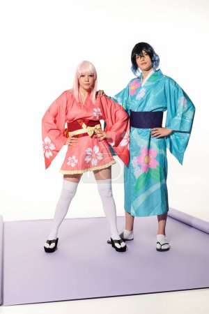 Foto de Pareja expresiva con confianza en coloridos kimonos y alas posando con las manos en las caderas, tendencia cosplay - Imagen libre de derechos