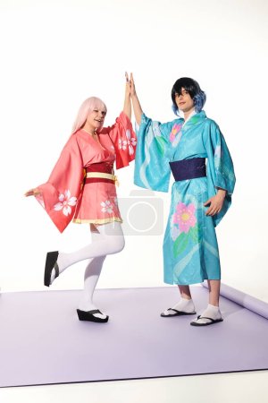 glückliche Cosplayer in leuchtendem Kimono und Perücken geben High Five auf lila Teppich und weißem Hintergrund