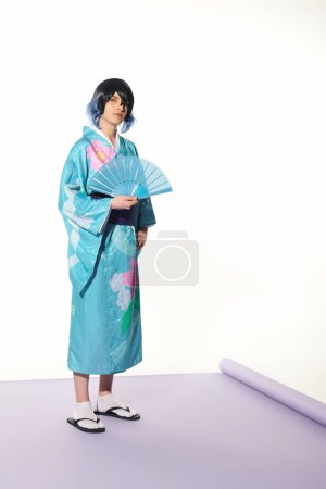 joven en kimono azul y peluca posando con abanico de mano sobre alfombra púrpura y fondo blanco, cosplay
