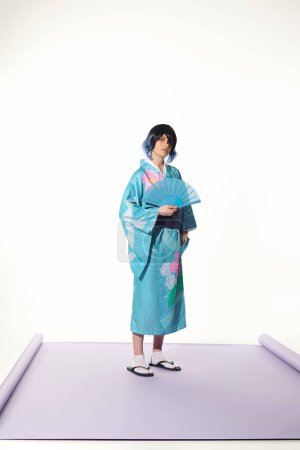 anime style man in blue kimono and wig holding hand fan and looking at camera on white backdrop