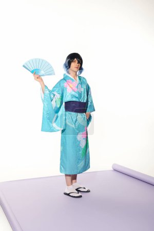 Cosplay-Stil, Mann in blauem Kimono und Perücke posiert mit Handfächer auf lila Teppich und weißem Hintergrund